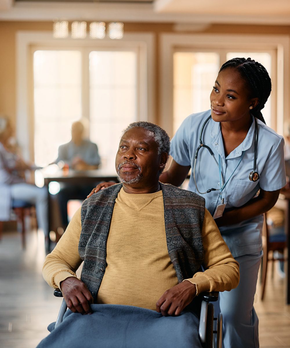 Black nurse and senior man in wheelchair looking through the window at nursing home. African American senior in wheelchair and young nurse looking through the window at residential care home.