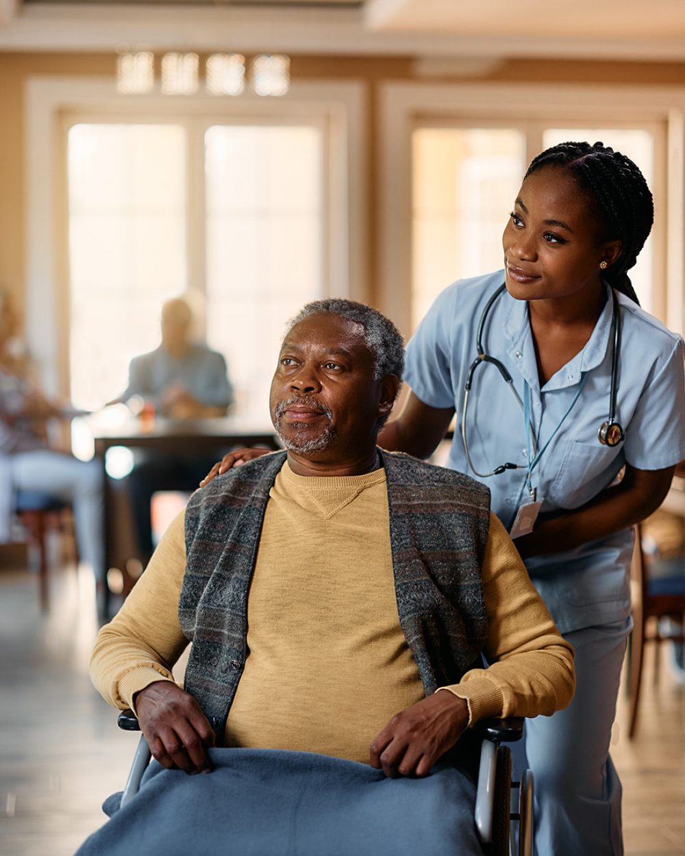 Black nurse and senior man in wheelchair looking through the window at nursing home. African American senior in wheelchair and young nurse looking through the window at residential care home.