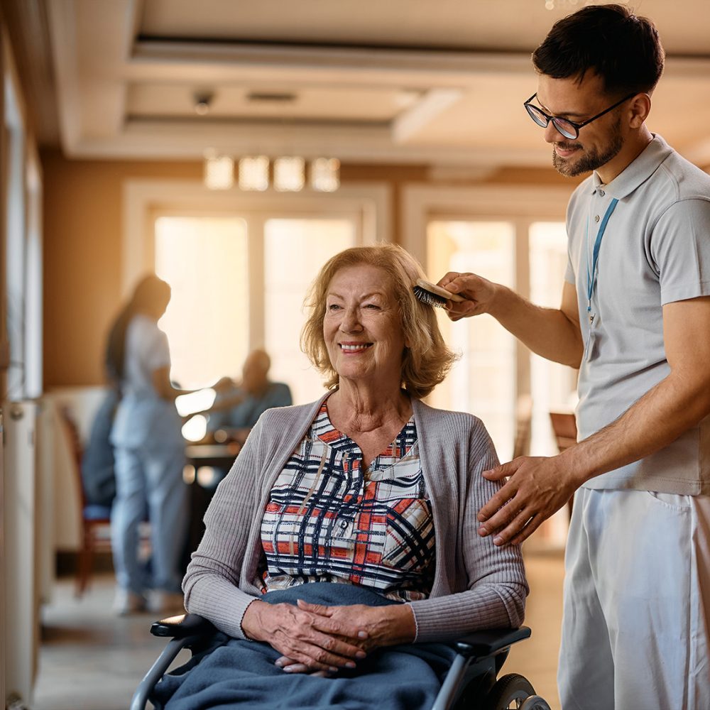 Happy caregiver brushing senior woman’s hair at nursing home. Happy senior woman in wheelchair enjoying while male nurse is brushing her hair at residential care home.