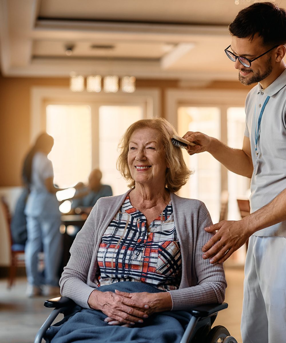 Happy caregiver brushing senior woman’s hair at nursing home. Happy senior woman in wheelchair enjoying while male nurse is brushing her hair at residential care home.